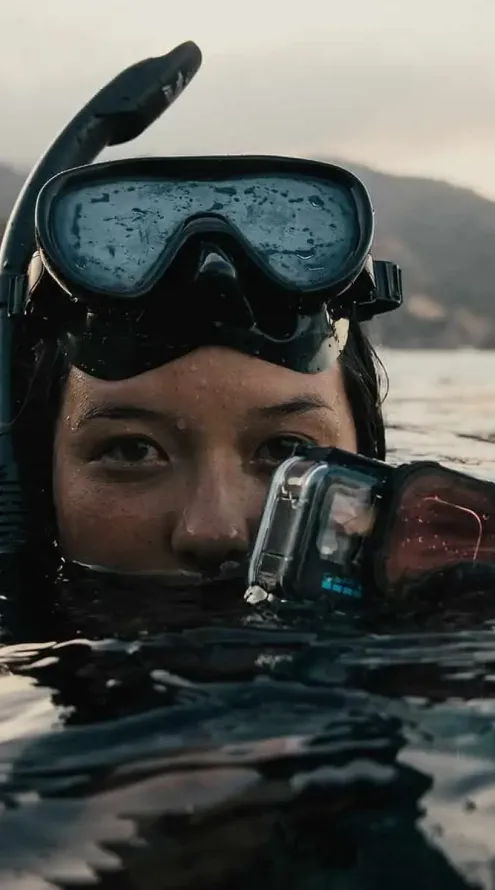 Person wearing a snorkel mask and holding an action camera while floating on the water surface.
